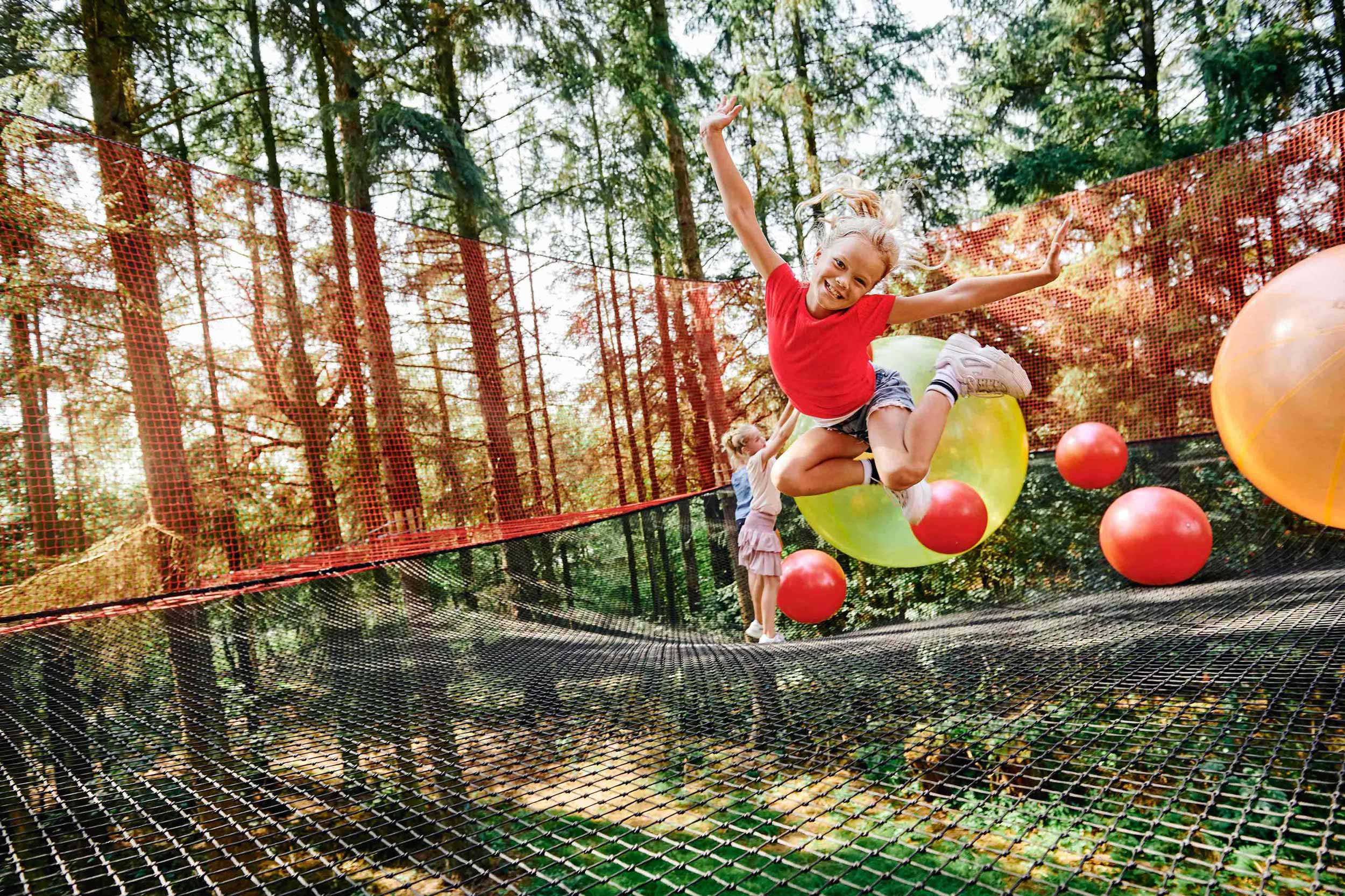 Børn leger i skoven i et stort net. Solen skinner. En pige springer op i luften og ser glad ud.  