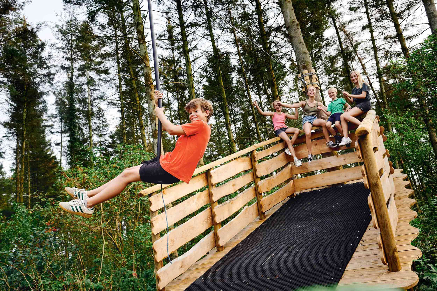 A boy is trying a zip line. He looks happy. There are four other children behind him, watching him.