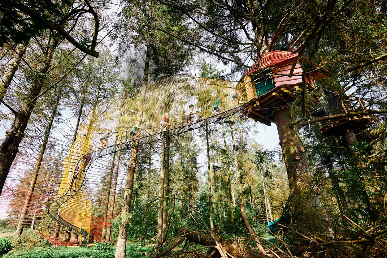 A long net bridge in beautiful colours leads up to a treetop house. Children are on their way up to the house.