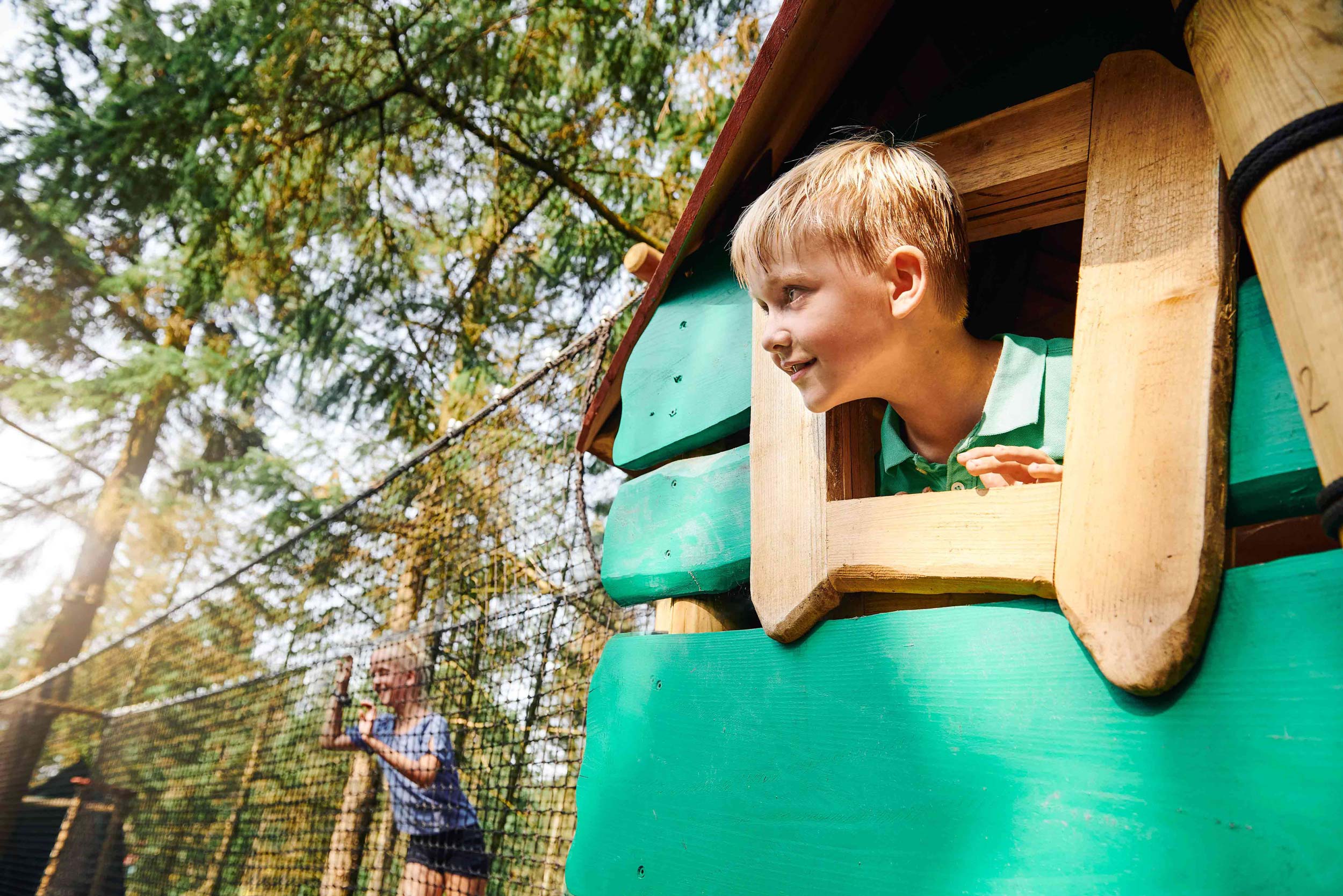 A boy is looking out of a hole (a window without glass) in a treetop hut.