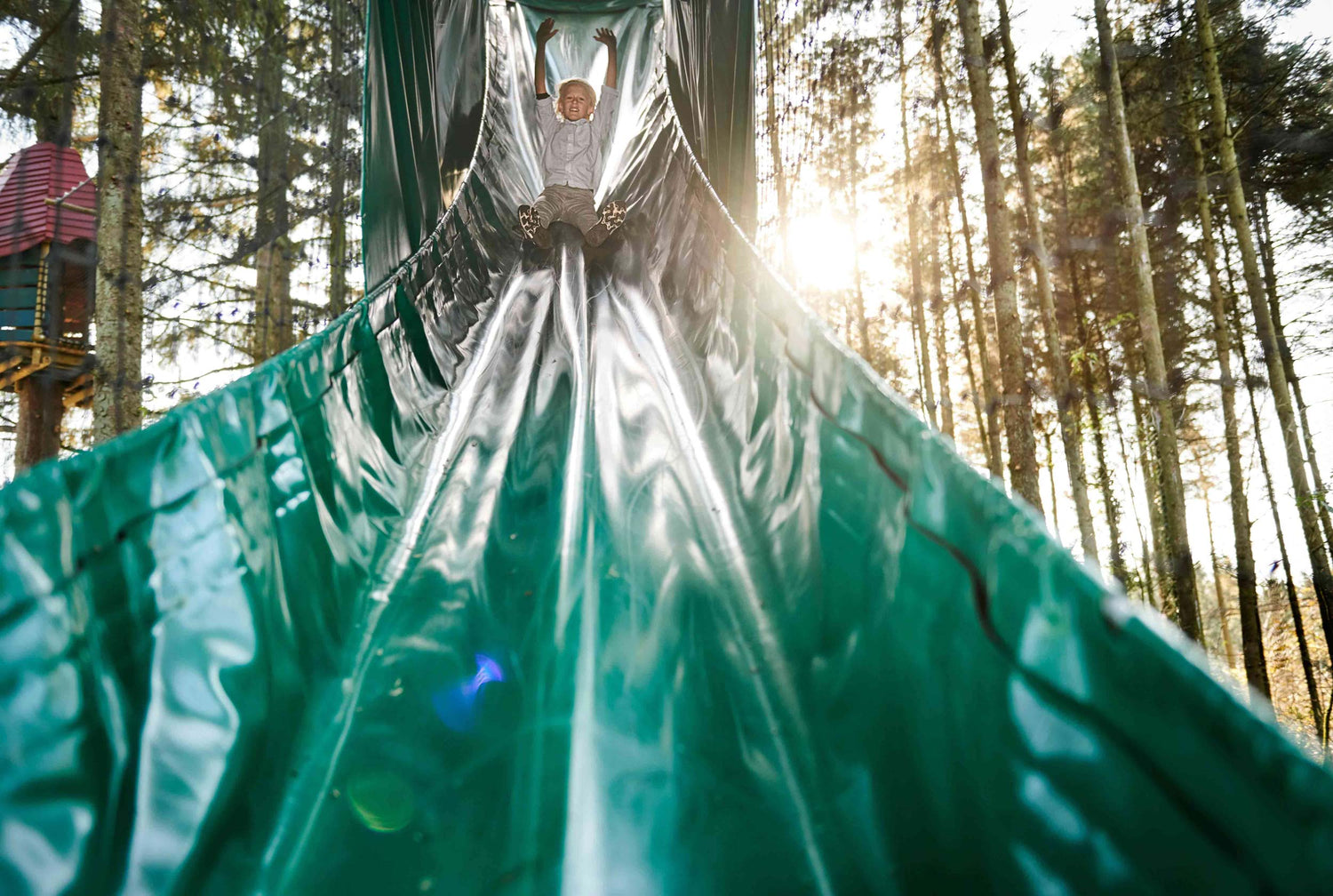 A boy is going down a green slide in WOW PARK, while he raises his arms in the air.